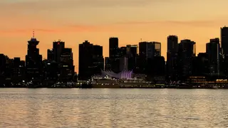 A dark waterfront skyline rises behind a lit sail-like structure and rippling water beneath an orange sunset sky.