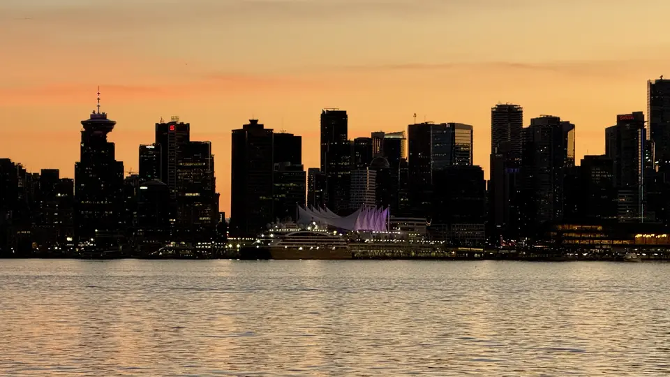 A dark waterfront skyline rises behind a lit sail-like structure and rippling water beneath an orange sunset sky.