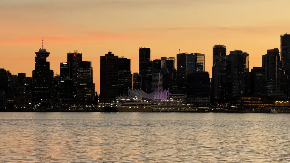 A dark waterfront skyline of high-rise buildings stands behind rippling water beneath an orange sunset sky.
