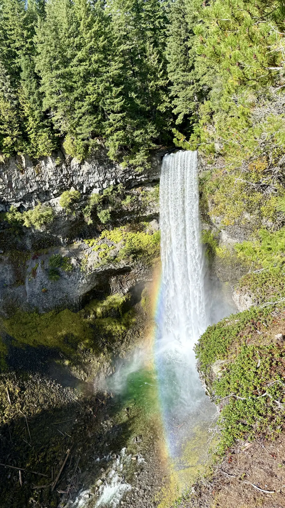 A tall waterfall drops over a rocky cliff into a forested gorge, with a rainbow glowing in the mist below.