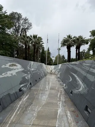 A wet concrete walkway runs between sloping gray walls beneath palm trees and flagpoles, with a domed building visible in the distance.