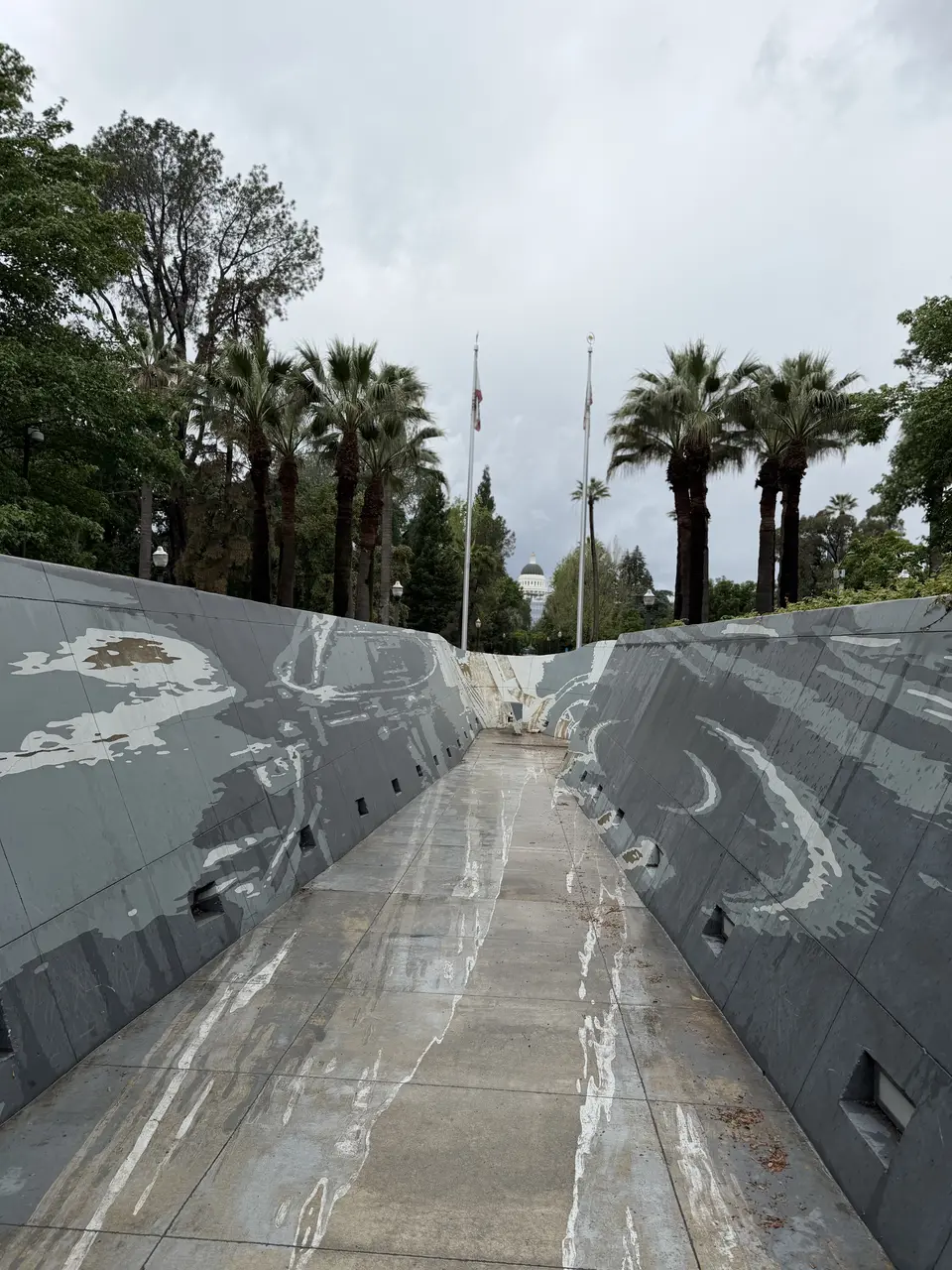 A wet concrete walkway runs between sloping gray walls beneath palm trees and flagpoles, with a domed building visible in the distance.