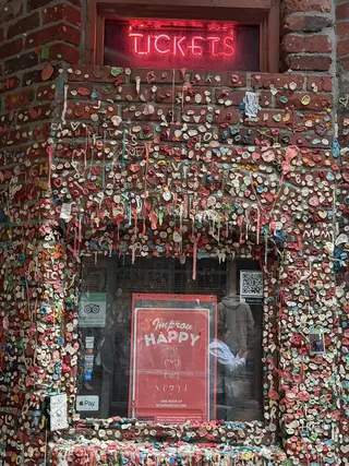 A red neon "TICKETS" sign glows above a window and brick wall densely covered with multicolored chewing gum.