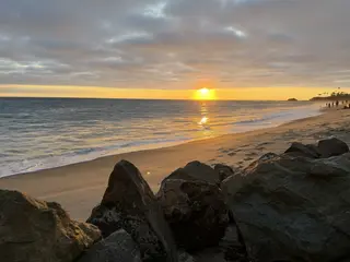 A golden sun sets over a sandy beach with rocks in the foreground.