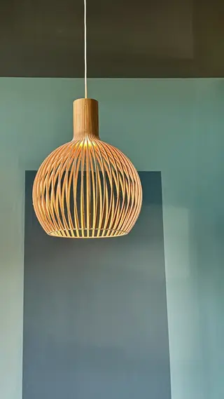A glowing woven wooden pendant lamp hangs against a teal wall above a dark rectangular panel.