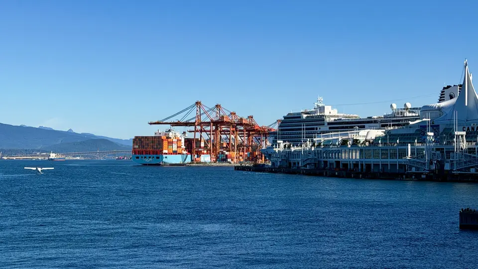 Canada Place and a cruise ship stand beside container cranes and a cargo vessel on Burrard Inlet beneath a clear blue sky.