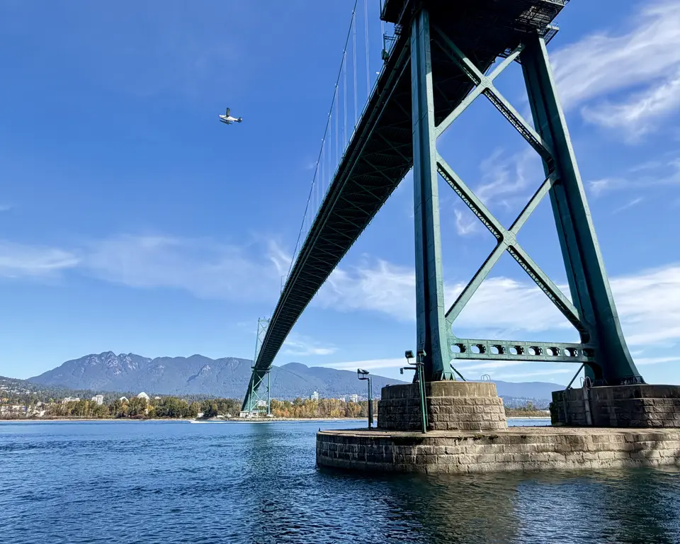 The Lions Gate Bridge spans blue water beneath a small airplane, with forested mountains in the distance.