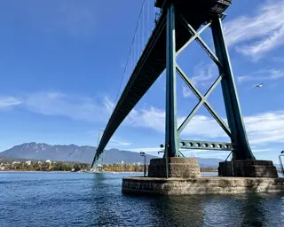 The Lions Gate Bridge spans blue water beneath a bright sky, with mountains and a shoreline in the distance.