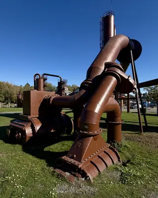 Rust-colored industrial pipes and machinery stand on a grassy lawn beneath a clear blue sky.
